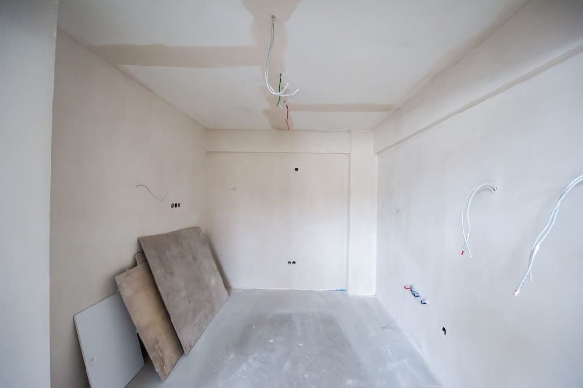 Empty room under renovation, with smooth unpainted walls and ceiling, loose cables coming out of the wall where the sockets will go and a few boards leaning in a corner.