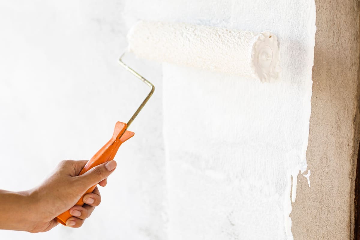 Mano con un rodillo extendiendo pintura blanca sobre una pared; a la derecha queda una franja de enlucido todavía sin pintar.