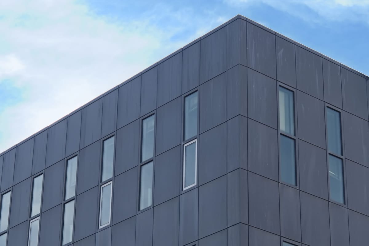 Corner of a multi-storey building clad in large grey panels separated by thin joints, with narrow windows and blue sky behind.