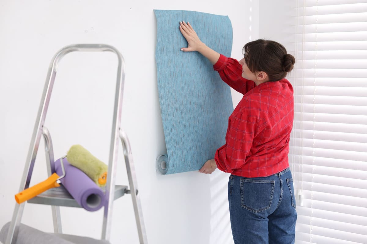 Someone hanging a strip of light blue wallpaper on a room wall, with a stepladder and paint rollers nearby.