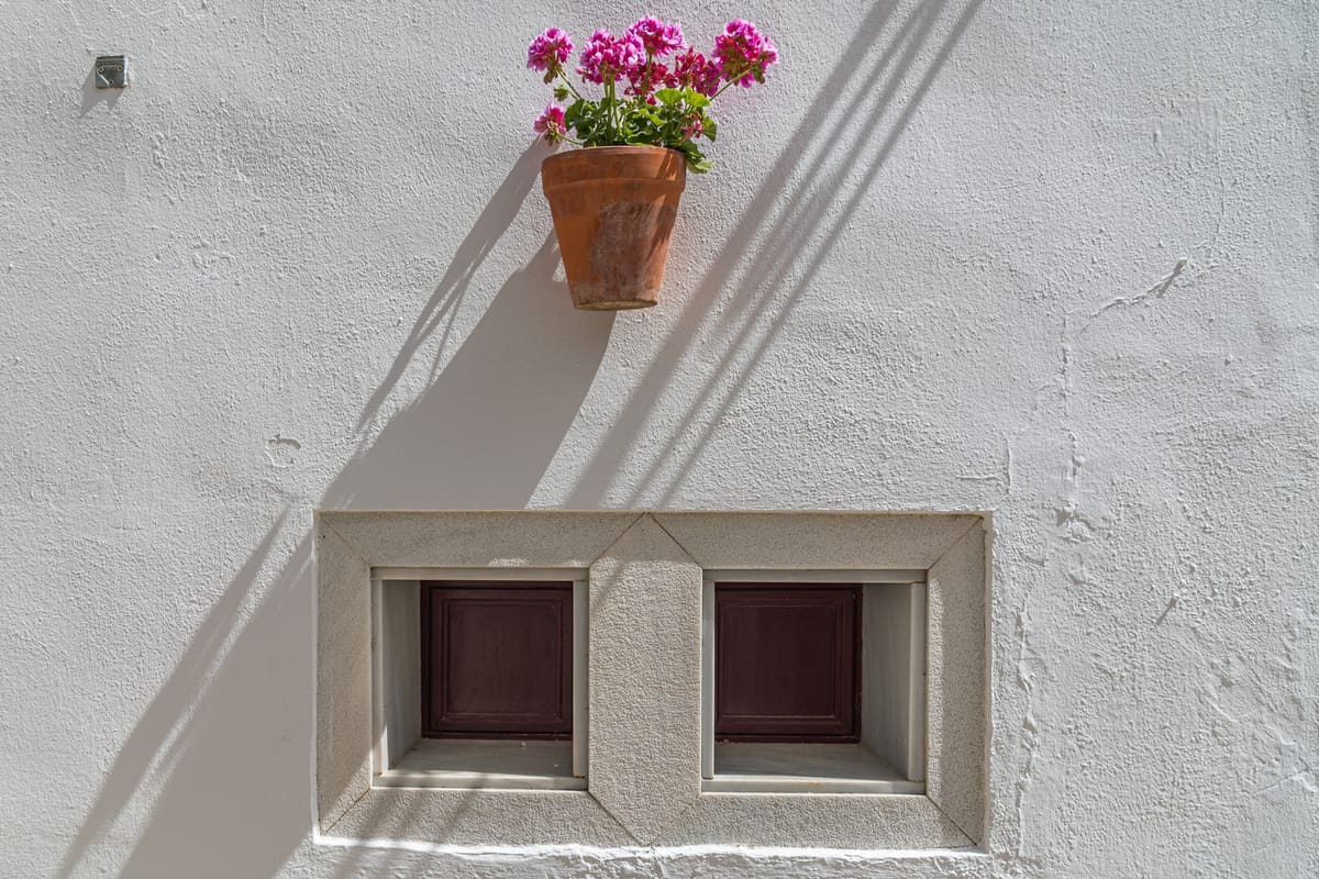 A limewashed wall on a street in Arcos de la Frontera, with a pot of geraniums hanging above a small double window. The layers of lime and a few fine cracks are visible.