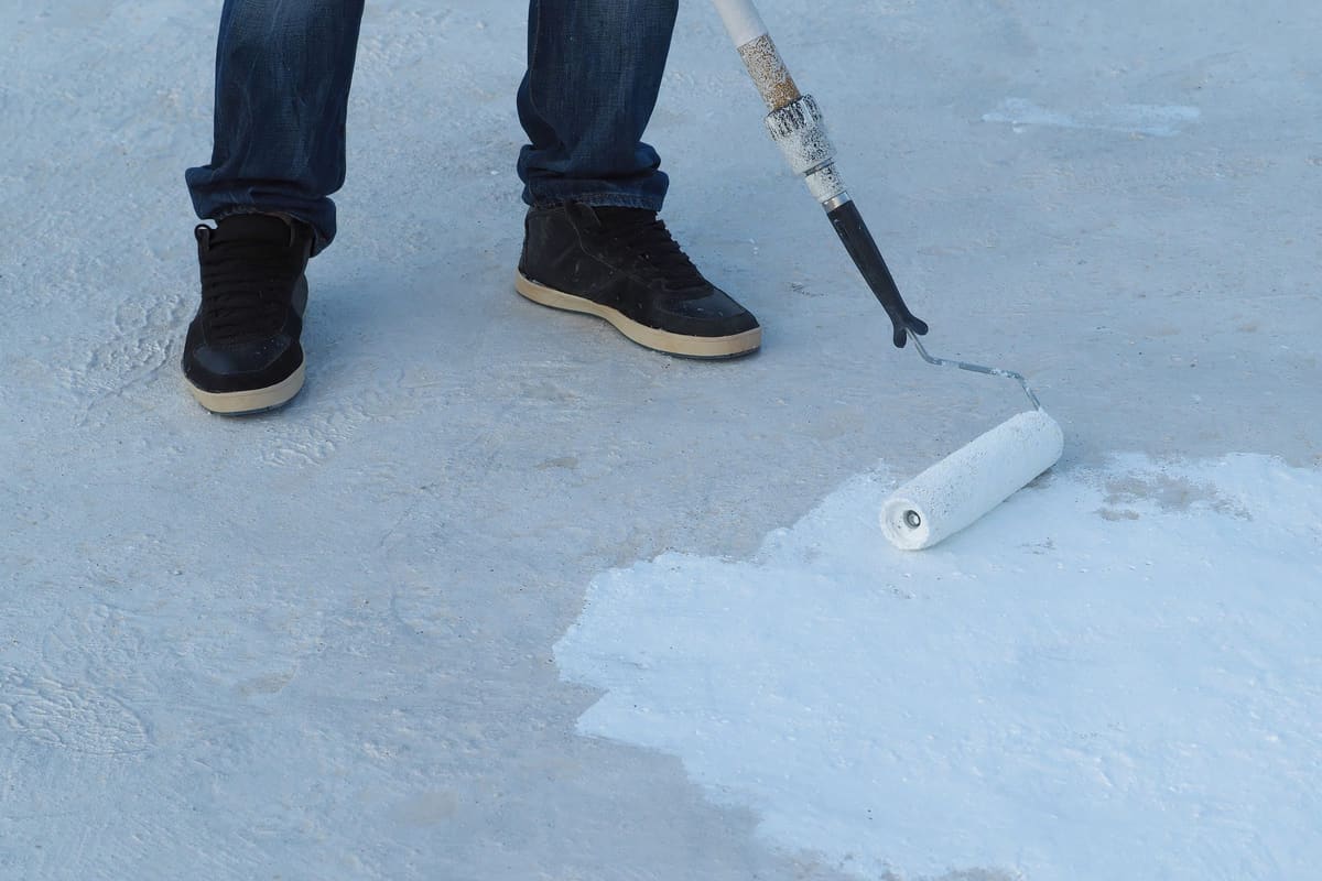 A roller spreading a coat of white paint over the grey concrete of a flat roof, beside the feet of the person using it.