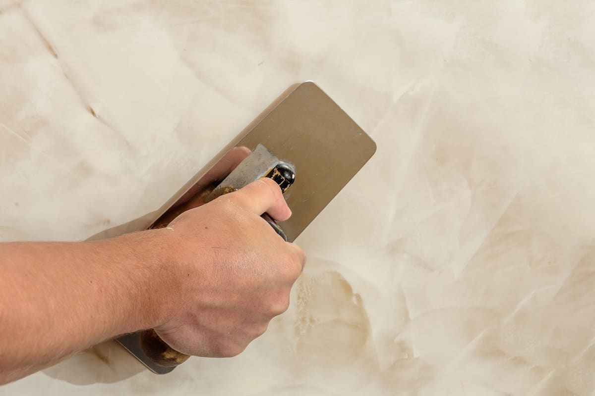 Mano pasando una llana de acero sobre una pared de estuco veneciano en tonos crema, con aguas más claras y más oscuras.