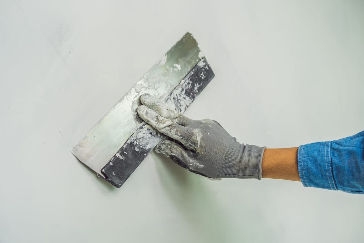 Mano con guante extendiendo una capa fina de pasta de alisar con una espátula ancha sobre una pared.